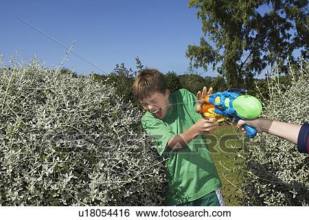 Stock Photograph - Two boys (6-11) playing with water pistols among bushes laughing close-up of hand with gun. Fotosearch