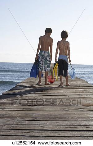 Two boys (6-11) walking on jetty carrying fishing nets and swim fins back view View Large Photo Image Picture - Two boys (6-11) walking on jetty carrying fishing nets and swim fins back view. Fotosearch