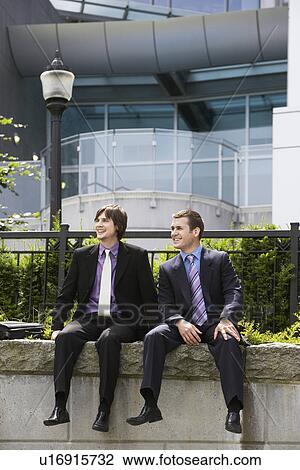 Stock Image - Two business men sitting on wall outside office. Fotosearch
