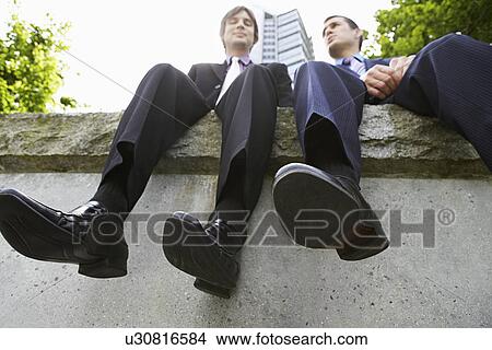 Two business men sitting side by side on wall low angle view View Large Photo Image Picture - Two business men sitting side by side on wall low angle view. Fotosearch