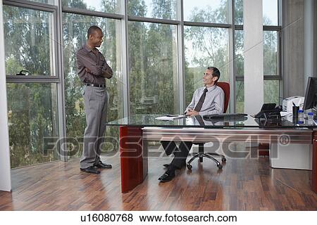 Two business men talking at desk in office View Large Photo Image Stock Photo - Two business men talking at desk in office. Fotosearch