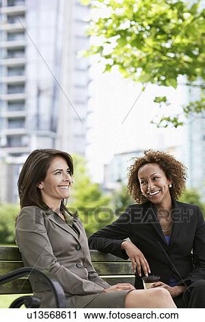 Stock Image - Two business women sitting on park bench smiling. Fotosearch