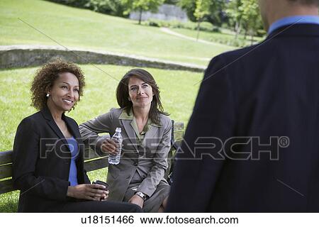 Stock Photograph - Two business women sitting on park bench looking at businessman. Fotosearch