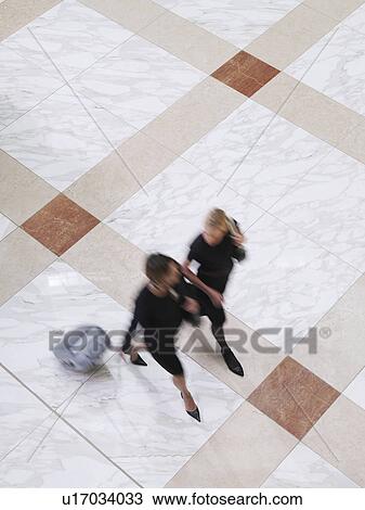 Stock Image - Two business women walking with suitcase elevated view long exposure. Fotosearch