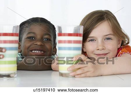 Stock Image - Two girls (5-6) leaning on table holding colourful glasses portrait close-up. Fotosearch