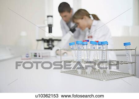 Stock Image - Two scientists using microscope in laboratory focus on test tubes in foreground. Fotosearch