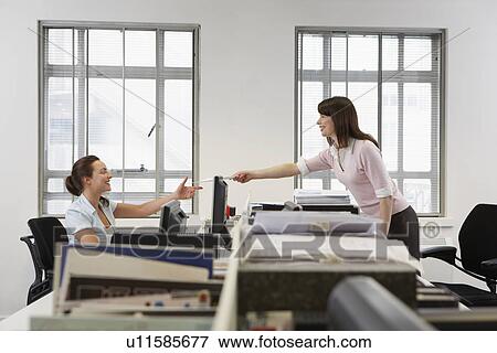 Stock Photo - Two women in office one handing documents to other over desk side view. Fotosearch