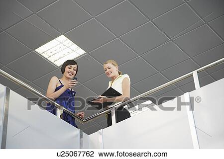 Stock Image - Two women looking over railing of indoor balcony. Fotosearch