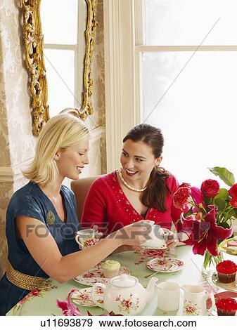Stock Photo - Two young women sitting at dining table pouring tea elevated view. Fotosearch
