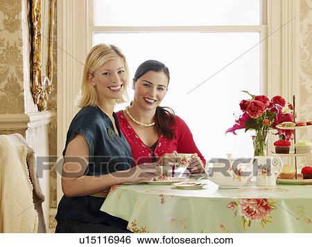 Stock Photograph - Two young women sitting at dining table drinking tea portrait. Fotosearch