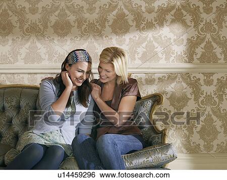 Stock Photograph - Two young women sitting on sofa smiling. Fotosearch