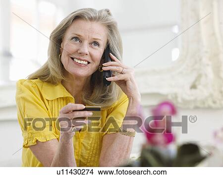 Picture - Woman holding credit card using phone in living room portrait. Fotosearch