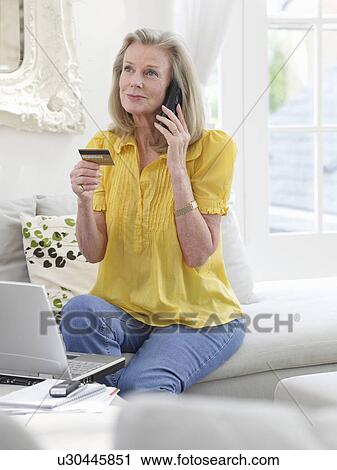 Stock Image - Woman holding credit card using phone in living room. Fotosearch