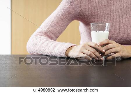 Stock Image - Woman holding glass of milk on table mid section. Fotosearch