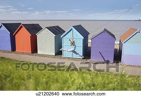 Stock Image - Woman jumping in front of beach hut. Fotosearch