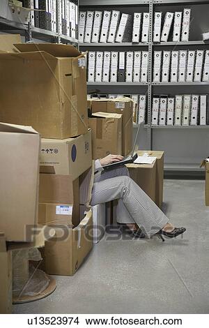 Picture - Woman using laptop sitting on floor between boxes in storage room low section. Fotosearch