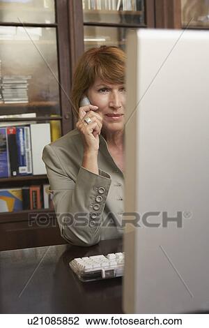 Stock Image - Woman using mobile phone while on computer in living room. Fotosearch