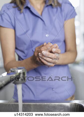 Stock Image - Woman washing hands in kitchen sink mid section. Fotosearch