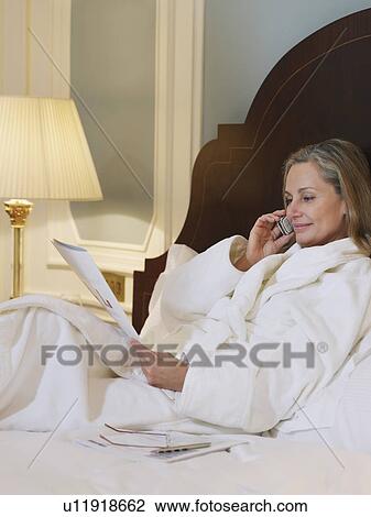 Stock Image - Woman wearing bathrobe using mobile phone lying on bed. Fotosearch
