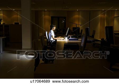 Stock Photo - Woman working at laptop in dark office. Fotosearch