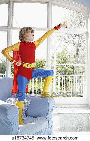 Stock Photography - Young boy (7-9) wearing superman costume standing on armchair and pointing. Fotosearch