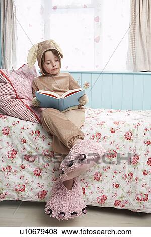 Stock Photo - Young girl (5-6) sitting on bed wearing bunny costume and monster slippers reading book. Fotosearch