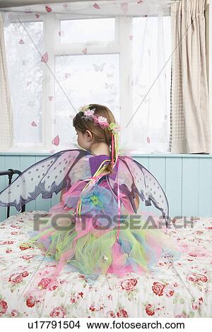 Young girl (5-6) wearing fairy costume sitting on bed by window back view View Large Photo Image Picture - Young girl (5-6) wearing fairy costume sitting on bed by window back view. Fotosearch