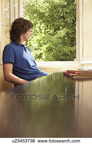 Stock Photo - Young man looking out of living room window. Fotosearch