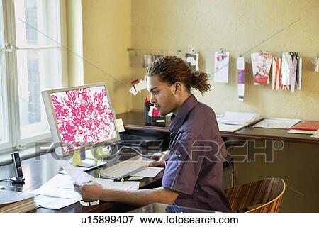 Young man reading papers near computer in office View Large Photo Image Stock Photo - Young man reading papers near computer in office. Fotosearch