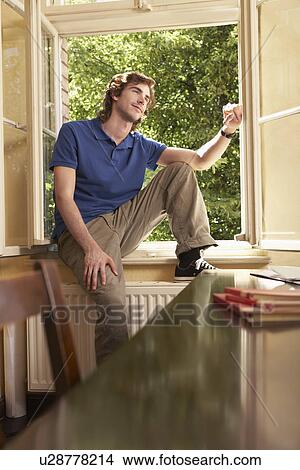 Young man sitting on sill of living room window View Large Photo Image Picture - Young man sitting on sill of living room window. Fotosearch