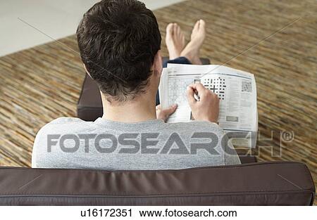 Young man sitting on sofa doing crossword puzzle in newspaper back view View Large Photo Image Stock Image - Young man sitting on sofa doing crossword puzzle in newspaper back view. Fotosearch