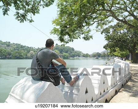 Young man sitting on surrounding wall by river back view View Large Photo Image Picture - Young man sitting on surrounding wall by river back view. Fotosearch
