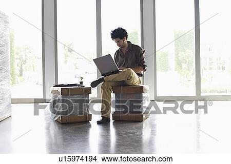Young man using laptop sitting on packages in empty office View Large Photo Image Picture - Young man using laptop sitting on packages in empty office. Fotosearch