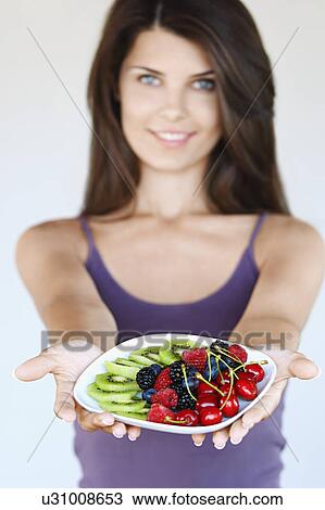 Stock Image - Young woman holding plate of fruit focus on foreground. Fotosearch