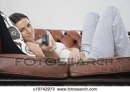 Stock Image - Young woman holding remote control lying on sofa. Fotosearch
