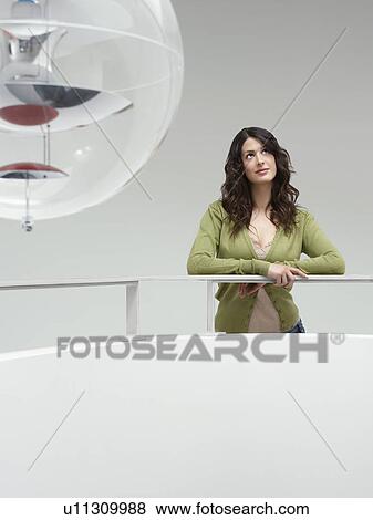 Stock Photo - Young woman leaning on railing indoors looking up. Fotosearch