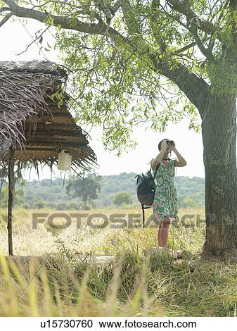 Stock Image - Young woman looking through binoculars by thatched roof. Fotosearch