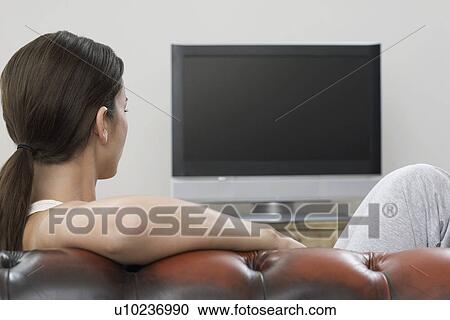 Stock Image - Young woman on sofa watching flat screen television. Fotosearch