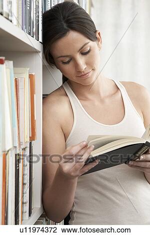 Young woman reading book leaning against bookshelf View Large Photo Image Stock Image - Young woman reading book leaning against bookshelf. Fotosearch