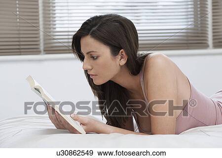Stock Photo - Young woman reading book lying on bed. Fotosearch