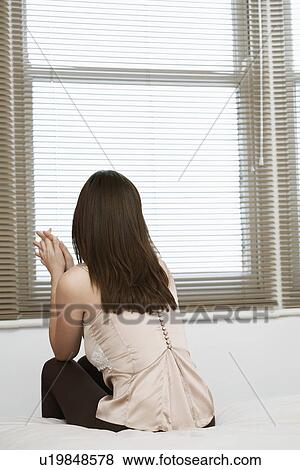 Stock Photo - Young woman sitting on bed by window back view. Fotosearch