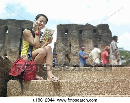 Young woman sitting on steps talking on mobile phone holding guide book View Large Photo Image Picture - Young woman sitting on steps talking on mobile phone holding guide book. Fotosearch