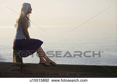 Stock Image - Young woman sitting on wooden bale on beach side view. Fotosearch