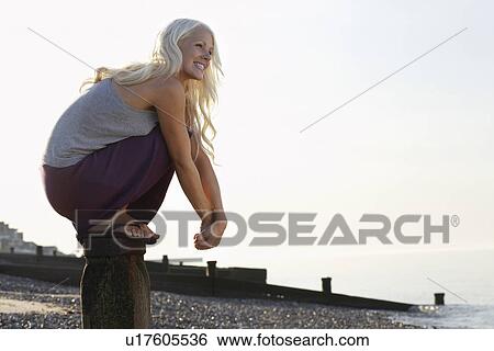 Stock Photograph - Young woman squatting on wooden pole on beach low angle view portrait. Fotosearch