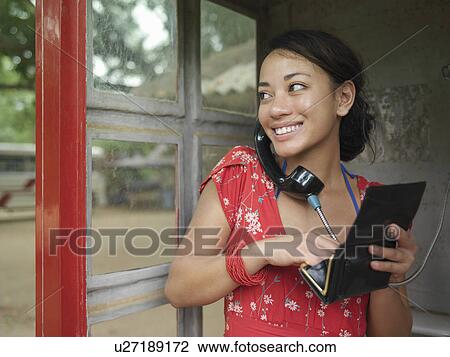 Stock Image - Young woman using public phone looking over shoulder holding purse smiling. Fotosearch