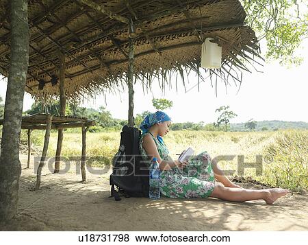 Stock Photo - Young woman wearing dress sitting in shade of hut roof. Fotosearch