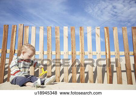 Stock Photo - Boy (1-2) playing with beach rake by wooden fence on sand dune. Fotosearch