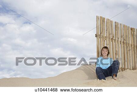 Stock Photo - Boy (10-12) sitting against wooden fence on sand dune portrait. Fotosearch