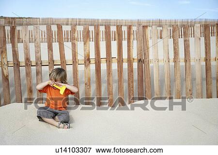 Boy (3-4) covering eyes sitting against wooden fence on sand dune View Large Photo Image Stock Photo - Boy (3-4) covering eyes sitting against wooden fence on sand dune. Fotosearch