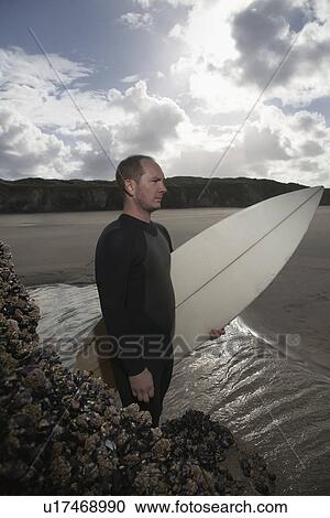 Stock Image - England Cornwall man holding surfboard standing by rocks on beach elevated view. Fotosearch